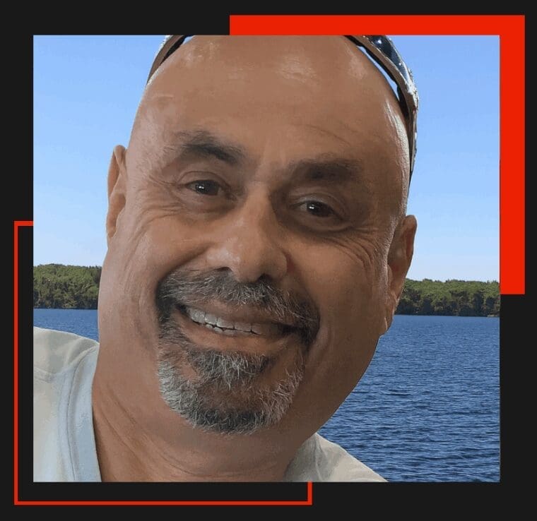 Man smiling by a lake, wearing sunglasses.